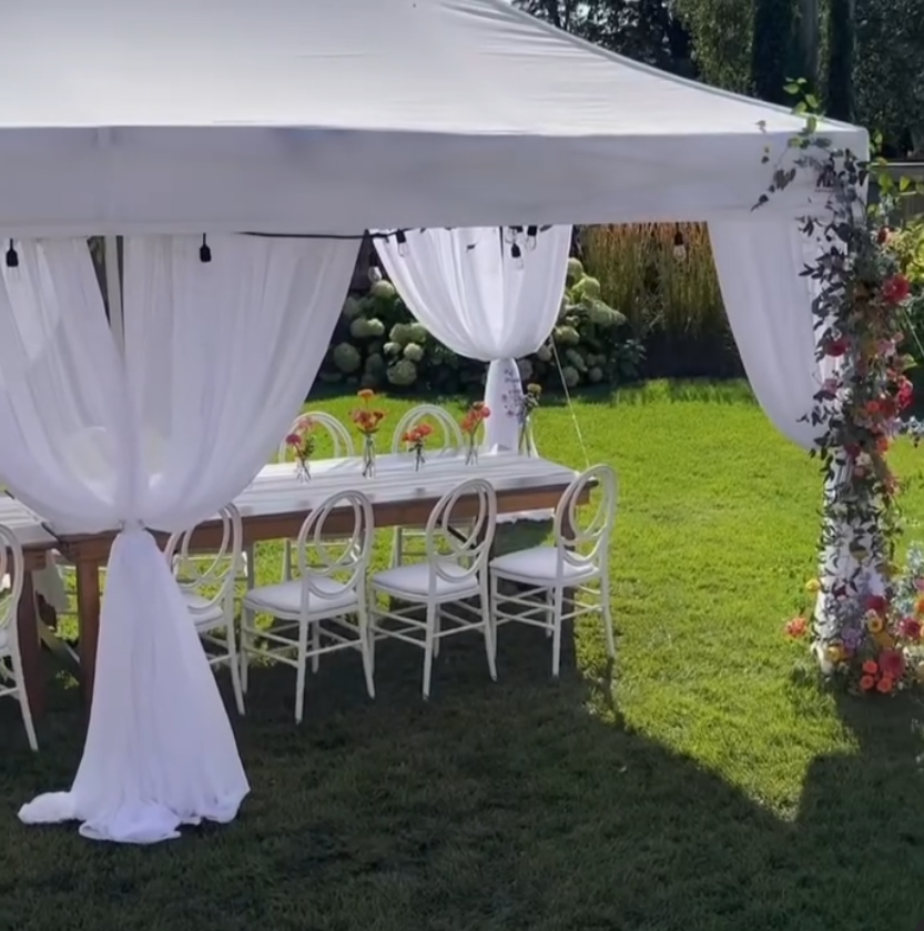 This image shows a white tent with white sheer drapes. Inside the tent, there is a beautiful table setting with chairs lined up neatly against the table